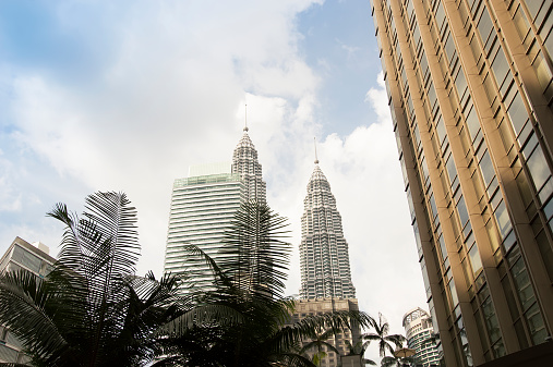 View of the Petronas Towers in Kuala Lumpur, Malaysia.  The view is from the bottom looking up.  There are several other buildings in the image on either side of the Petronas Towers.  There are also several trees and green ferns at the bottom.  The bright blue sky is lined with white clouds.