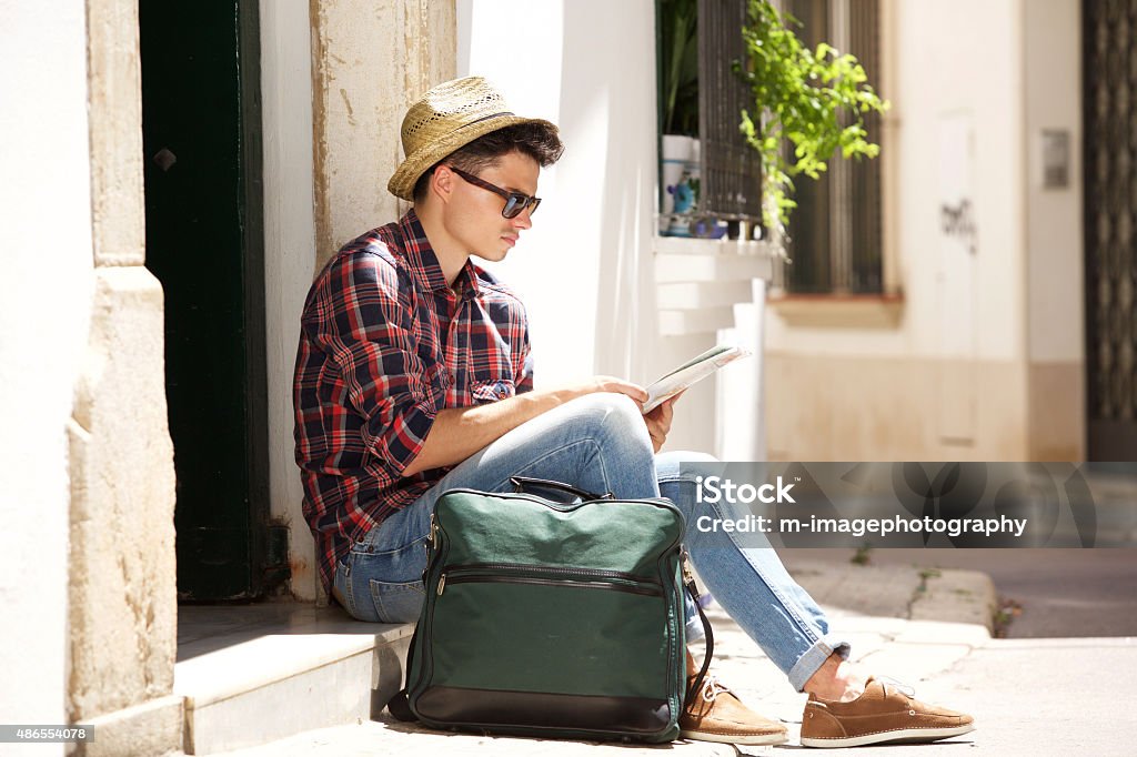 Traveling man sitting on sidewalk reading map Side portrait of a handsome young traveling man sitting on sidewalk reading map 20-29 Years Stock Photo