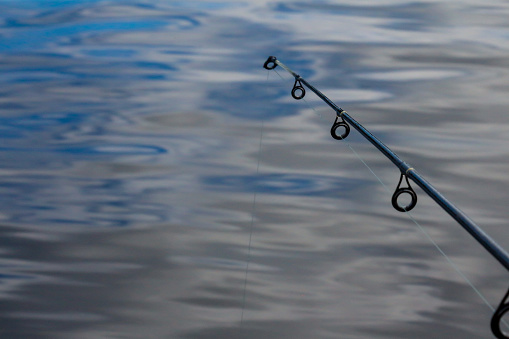 Image of a black fishing pole and clear fishing line in front of blue and grey water.  The image is a first person perspective from a person fishing in Ontario, Canada.