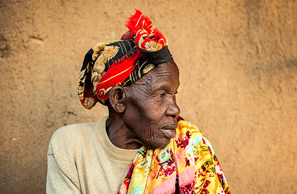 An old African woman sitting alone in her village, waiting for the sun to cool down so she can take the cows for a drink of water.