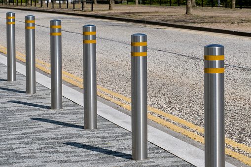 A row of bollards situated at the edge of a pavement next to a road. each Stainless Steel bollard has a reflective band