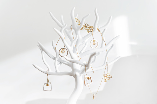 White jewelry display, stand hanger in the shape of tree with gold jewelery is standing on the shelf.