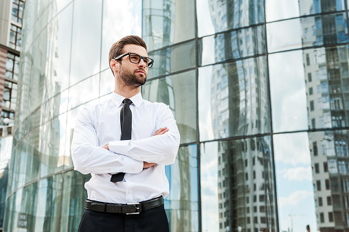 Low angle view of confident young businessman keeping arms crossed and looking away while standing outdoors with office building in the background