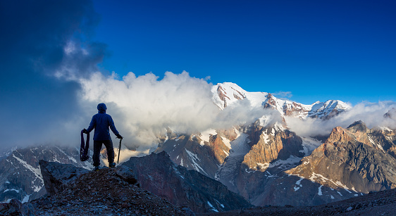 Silhouette Man Staying on Top of Rock Cliff Holding Climbing Gear Stormy Clouds and Peaks Illuminated bright Morning Sun