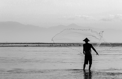 In Bali, Indonesia, a matured man wearing a straw hat throws his fishing net into the sea.