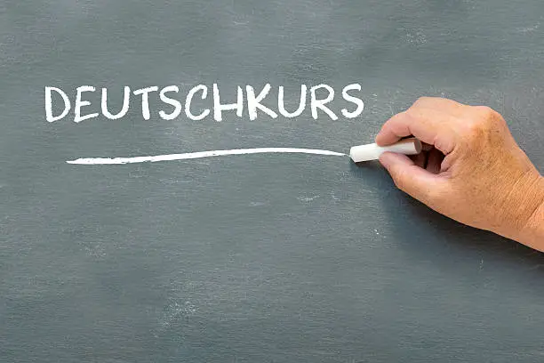 Hand on a chalkboard with the German word Deutschkurs (German course). Language class concept showing student hand writing on the blackboard.