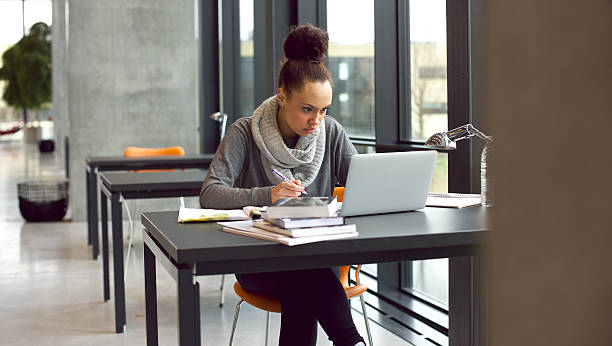 Young female student taking notes for her study Young afro american woman sitting at table with books and laptop for finding information. Young student taking notes from laptop and books for her study in library. exam student university writing stock pictures, royalty-free photos & images