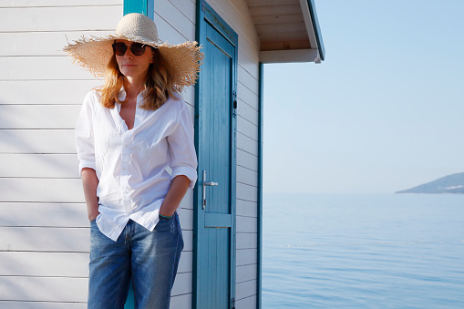 Portrait of smiling middle age woman standing on the beach. Beautiful female wearing straw hat and sunglasses while she is on holiday.