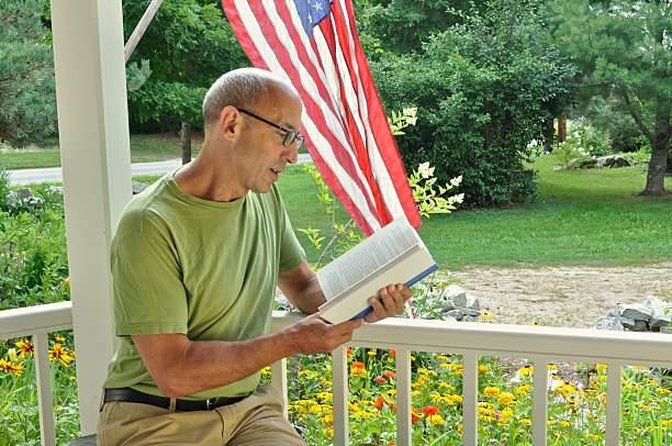 Senior Mature Man Reading Book on Porch with American Flag stock photo