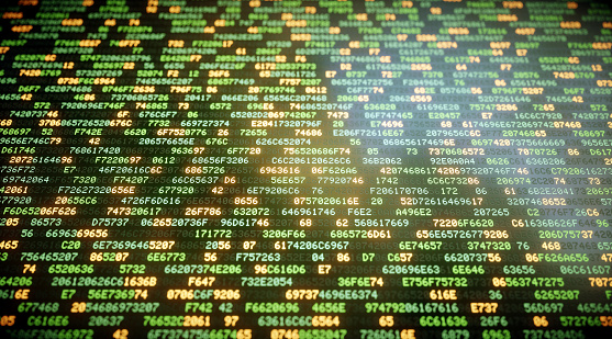 An abstract IT background concept representing encrypted data in hexadecimal format on a flat screen. Random blocks of data are organized in rows and come in a green hue. Some characters are glowing more brightly in a yellow hue. The screen surface reflects a glaring source of light. The image features a shallow depth of field.