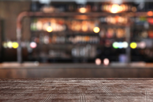 Close-up of a rough brown wooden table edge in a pub bar and cafeteria with defocused bar counter background. Daylight coming from the windows. Copy space on the table area and blurred image for easier editing and addition of graphic elements.