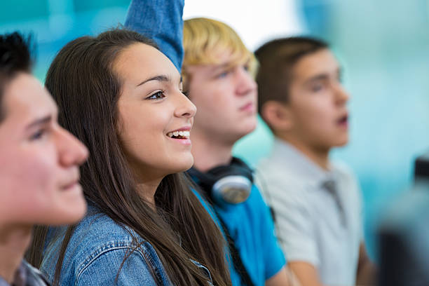 Teenage girl raising hand, asking question in high school class Teenage Hispanic girl is smiling and raising hand. She is answering or asking a question in high school classroomm. Girl is wearing a denim jacket. She is sitting at desk nex to diverse classmates. hand raised classroom student high school student stock pictures, royalty-free photos & images
