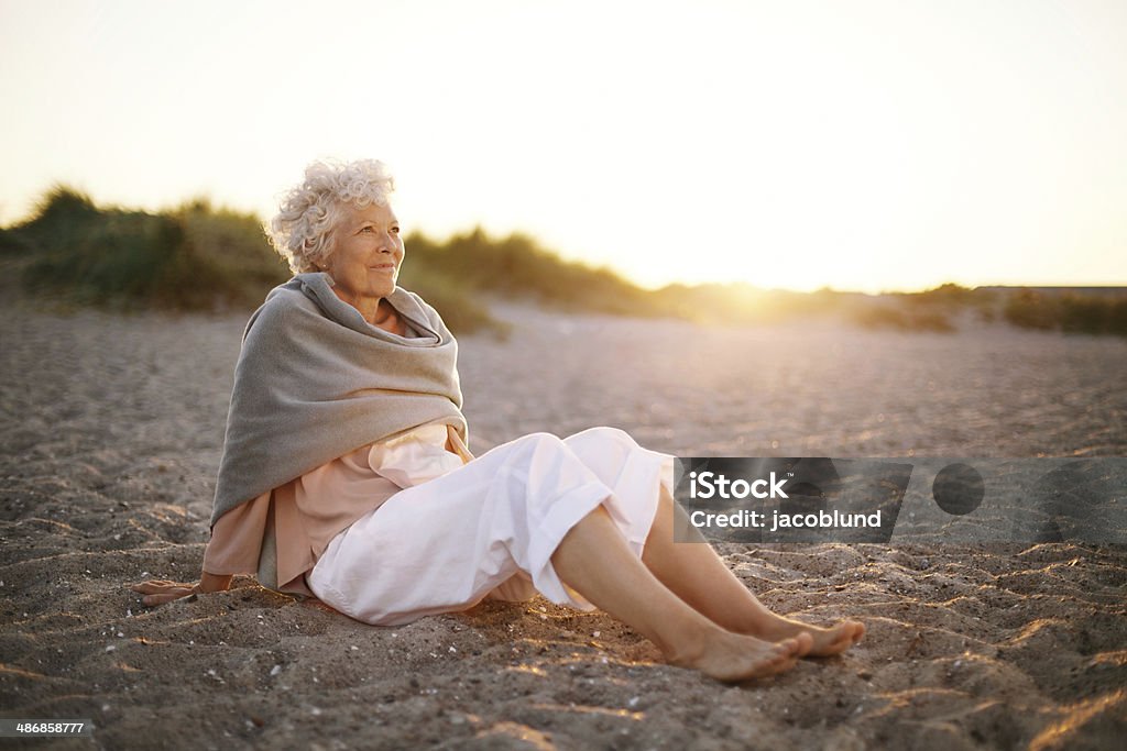 Relaxed elderly woman sitting on the beach Image of relaxed elderly woman sitting on the beach looking at a view. Senior woman wearing shawl sitting on beach. Mature Women Stock Photo