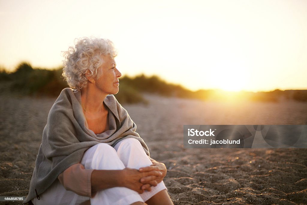 Mature woman on beach looking into horizon Old woman sitting on the beach looking away at copyspace. Senior female sitting outdoors Senior Adult Stock Photo