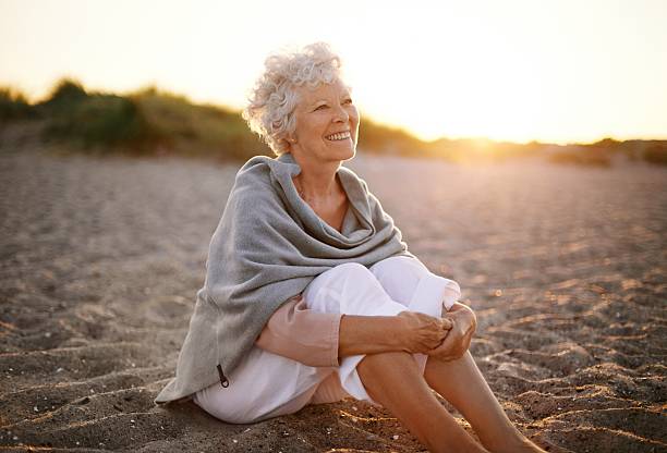 fröhlich alter frau sitzt auf dem strand - seniorinnen stock-fotos und bilder