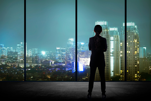 Woman standing in his office looking at the city at night. Business success concept