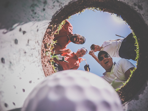 A Point Of View picture of 4 Golf Players and Golf Ball view From Inside the Hole on a sunny day of summer. The players are excited and happy.