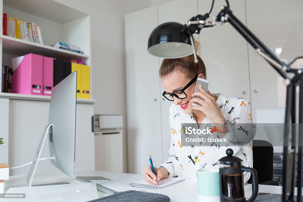 Woman working in an office, talking on smart phone Smiling blonde woman wearing nerd glasses sitting at the desk in an office and talking on smart phone, taking notes. Desk Stock Photo