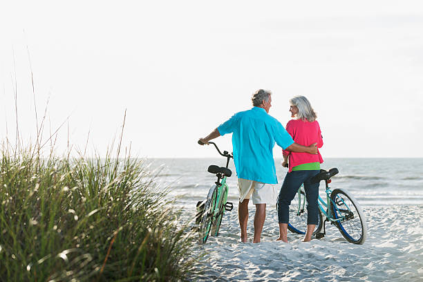matura coppia con biciclette in spiaggia - mature adult bicycle senior adult heterosexual couple foto e immagini stock