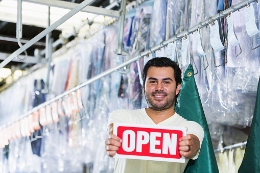 Portrait of successful Hispanic small business owner of a dry cleaning business, holding an OPEN sign.  He is standing in front of racks of clothing on hangers, covered in plastic, smiling at the camera.