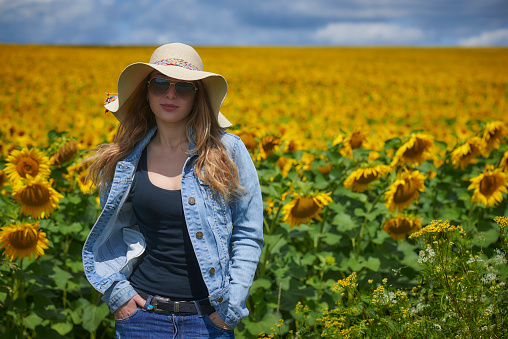 Attractive young blonde woman in a blue denim jacket, jeans and hat standing in sunflowers background. look at camera