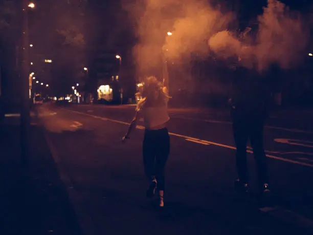 Photo of Teenagers running down city street with smoke flares at night