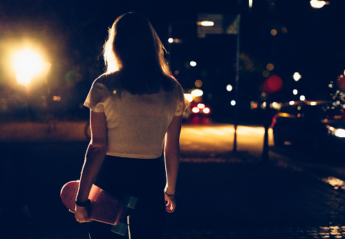 Rearview image of a skater girl walking at night alone with her pink  skateboard on a city street