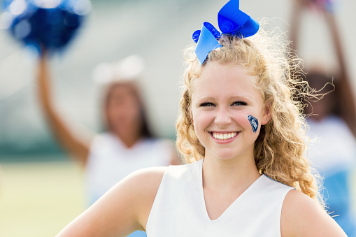 Pretty teenage Caucasian girl with blonde curly hair is smiling at camera. She is cheering during high school football game or cheer event. Girl is wearing blue hairbow and cheer uniform.