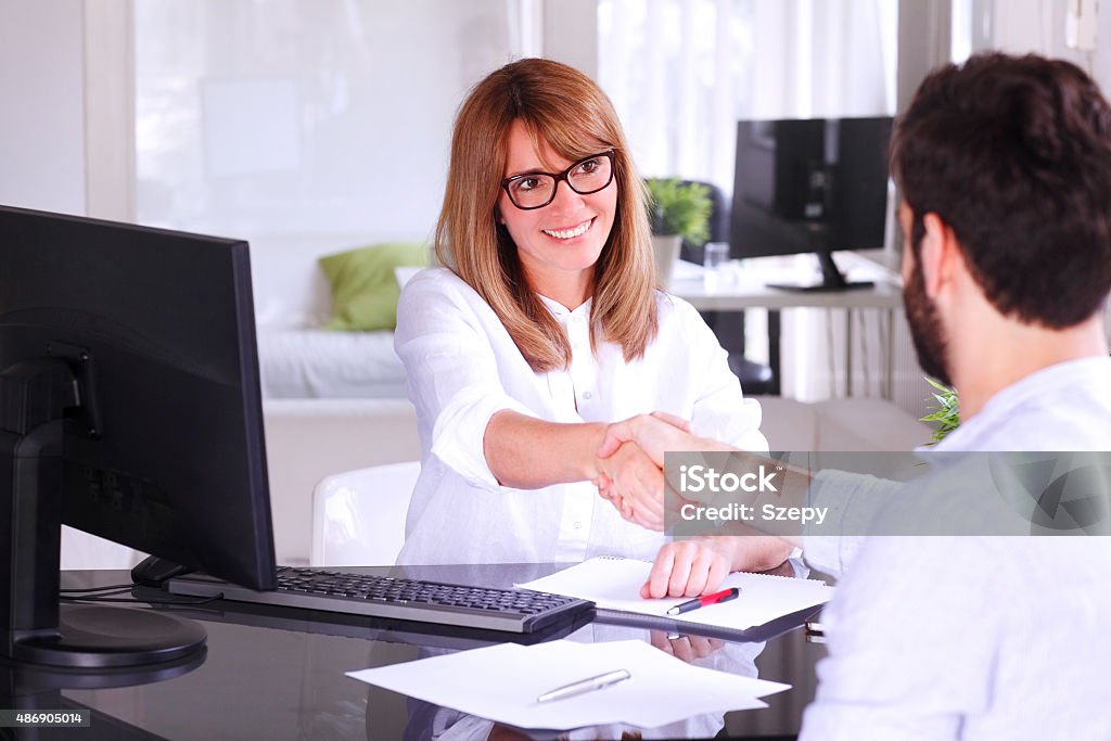 Shaking hands Portrait of business people shaking hands. Executive businesswoman sitting at office in front of computer and making successful business deal with young professional man. Handshake. 2015 Stock Photo