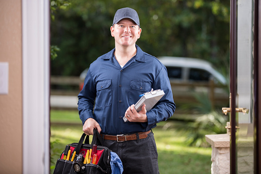 Caucasian repairman or blue collar/service industry worker makes service/house call at customer's front door. He holds his clipboard and tool box filled with work tools. Inspector, exterminator, electrician. He wears a navy blue uniform. Service truck seen in background parked on road. 