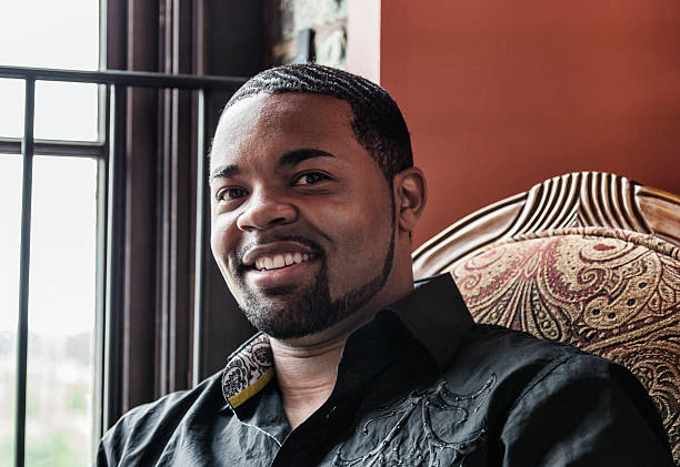 A handsome young black man wearing an embroidered black button down dress shirt is smiling and posing for a portrait while sitting in a comfortable, retro styled easy chair near a natural light window. Nashville, Tennessee iStock Sparkle & Shine Lypse - May 2012.