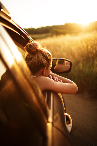 Young woman looking out of the window of her car and enjoys the view at sunset, getting away from the city 
