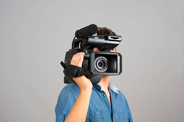 Cameraman holding a camera on his shoulder isolated on gray. Unrecognizable cameraman carrying hd camera on his shoulder. Man standing in front of gray background. Horizontal composition. Image taken in studio and developed from Raw format.