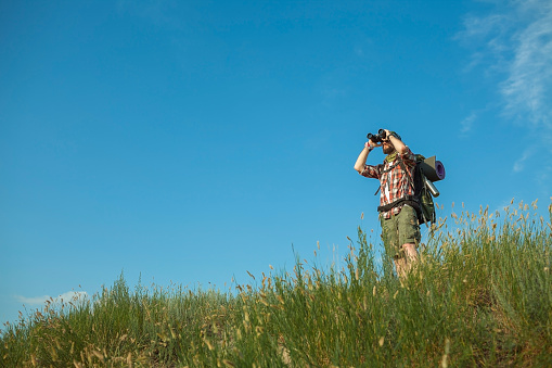 Young smilimg caucasian tourist with backpack  standing on the top of hill against blue sky background and looking through binoculars