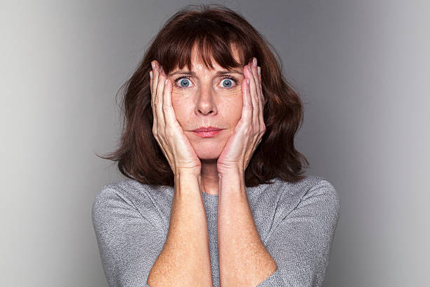 anxious senior woman under shock independent senior mature woman - portrait of anxious beautiful 50's woman under shock with both hands on face for fear and stress confused face stock pictures, royalty-free photos & images