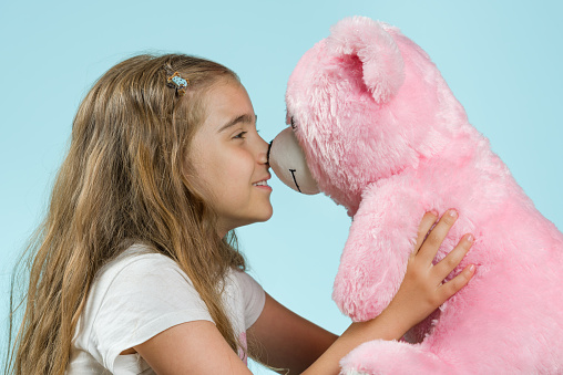 Happy girl smiles with her pink tedy bear. She holds and loves teddy bear and her eyes looks it. She is 8 years old. Positive human face expression. Isolated on light blue background. Studio shot.