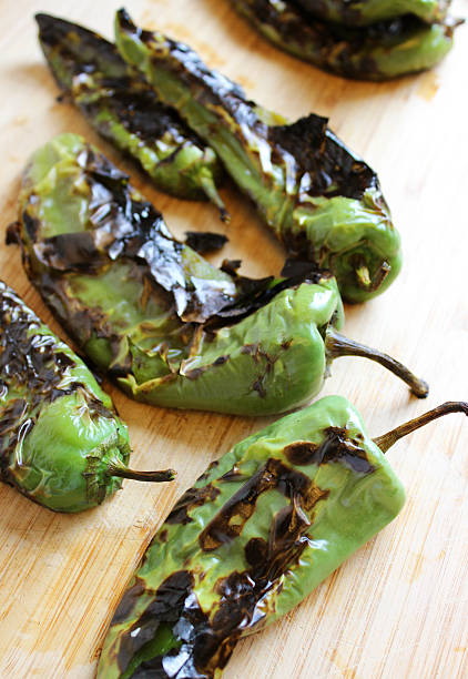 Roasted Blistered Green Chiles on Wooden Cutting Board A group or bunch of New Mexican green chiles.  They have just been roasted / blistered, as a part of their preparation for removing the stems, skins and seeds.  This process is necessary before using them to cook green chile sauce, stuffed chiles and any number of green chile recipes.  These green banana-shaped chile peppers go by names such as:  New Mexican green chile, Anaheim chile pepper, Big Jim, Sandia, or California green chile pepper.  The chiles rest on a wooden cutting board. anaheim pepper photos stock pictures, royalty-free photos & images