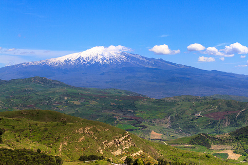 A tranquil, bucolic panorama of snowy Mt. Etna in spring, with blue sky and puffy clouds coming from the volcanic cone, and hilly green countryside in front. Shot from Troina (Enna Province). Copy space in the sky.