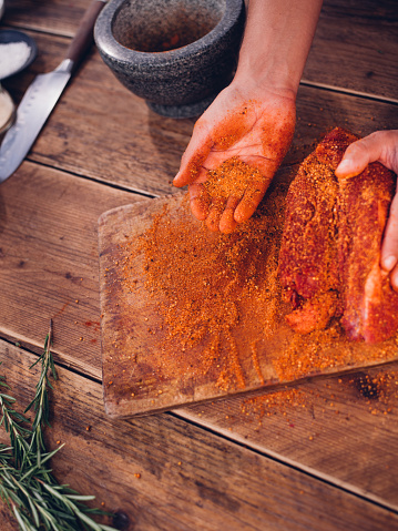 Cropped image of a person's hand showing some spicy seasoning on a wooden board being used for a dry meat rub to flavour a piece of raw pork