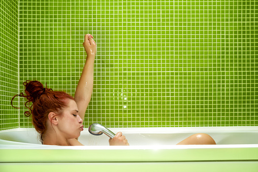 side view of woman in bathtub singing, enjoying bath and having fun.photo taken inside bathroom, green tile on the wall.