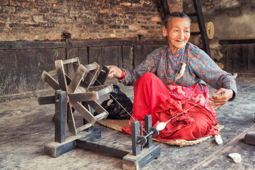 Kathmandu, Nepal - March 22, 2014: Old nepalese woman is spinning thread using spinning wheel in front of her house in Bhaktapur. Spinning is an ancient textile art in which plant, animal or synthetic fibers are drawn out and twisted together to form yarn. A spinning wheel is a device for spinning thread or yarn from natural or synthetic fibres.