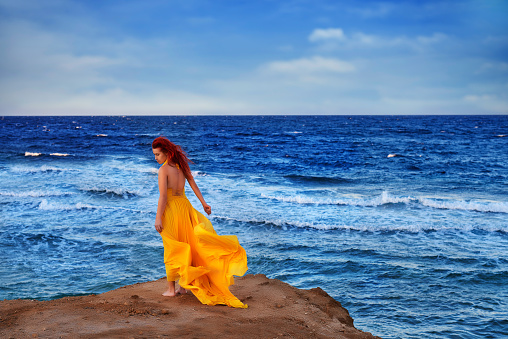 rear view of woman in yellow chiffon dress on beach enjoying tranquil day at sunset, sea and waves in background, fashionable look.