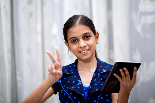 ten year girl playing with her tablet pc or tab in her house sitting on a sofa or a wooden chair against white curtain and showing a sign of victory