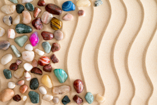 Arrangement of waterworn pebbles and colorful tumbled polished gemstones on golden beach sand with an undulating wavy pattern and copyspace