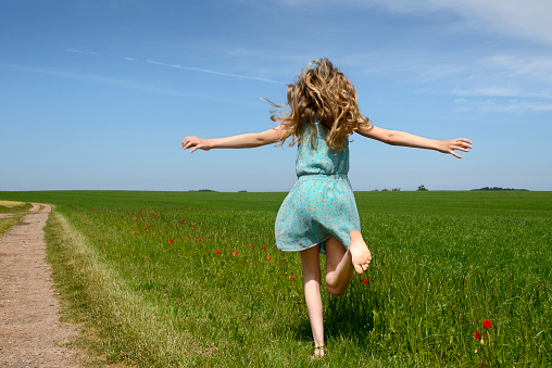 Teenage girl running on a green meadow with poppies under blue sky in springtime. The barefoot girl has long blond hair and she is wearing a light blue sundress. Her arms are outstretched. The girl has a northern european descent. Taken with copy space from rear view. Horizon over land in the background.