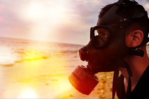 side view of man wearing gas mask, sitting at the edge of the sea and looking through polluated sea, toxic environment concept.