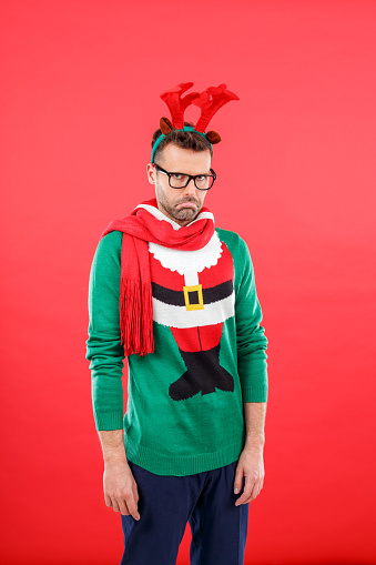 Studio portrait of sad man wearing santa christmas sweater and reindeer antlers headband, standing against red background and looking at camera.