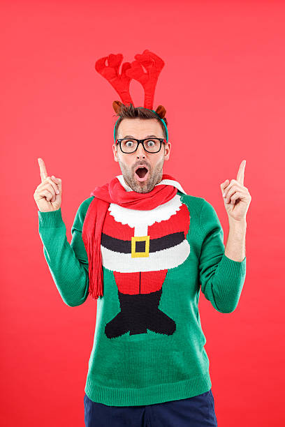Surprised nerd man in funny winter outfit against red background Studio portrait of surprised man wearing santa christmas sweater and reindeer antlers headband, standing against red background and pointing with index fingers at copy space, staring at camera with mouth open, rolling his eyes.  nerd sweater stock pictures, royalty-free photos & images