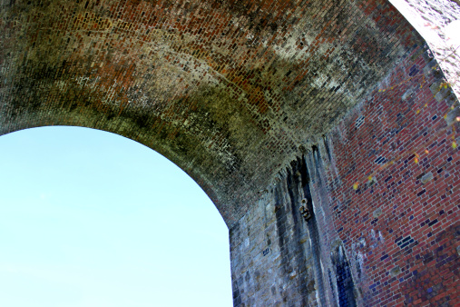 Photo showing the red brick architecture and curving underside of a large and rather weathered bridge / viaduct archway / arch that has been standing for many decades.