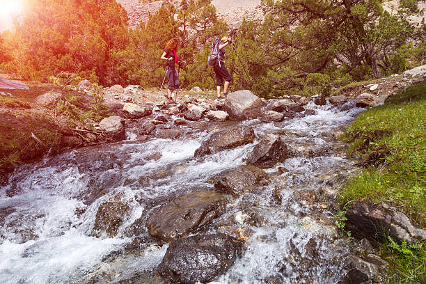 Two hikers crossing fast flowing river People going across mountain creek with fast streaming water jumping on the rocks green meadow and forest along river forest wild landscape and sun on background ford crossing stock pictures, royalty-free photos & images